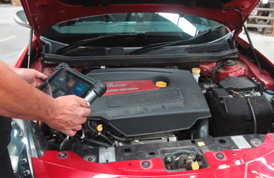 Photo of a man's hands holding ipad device that is showing readings from a car engine. Car engine is in the background.