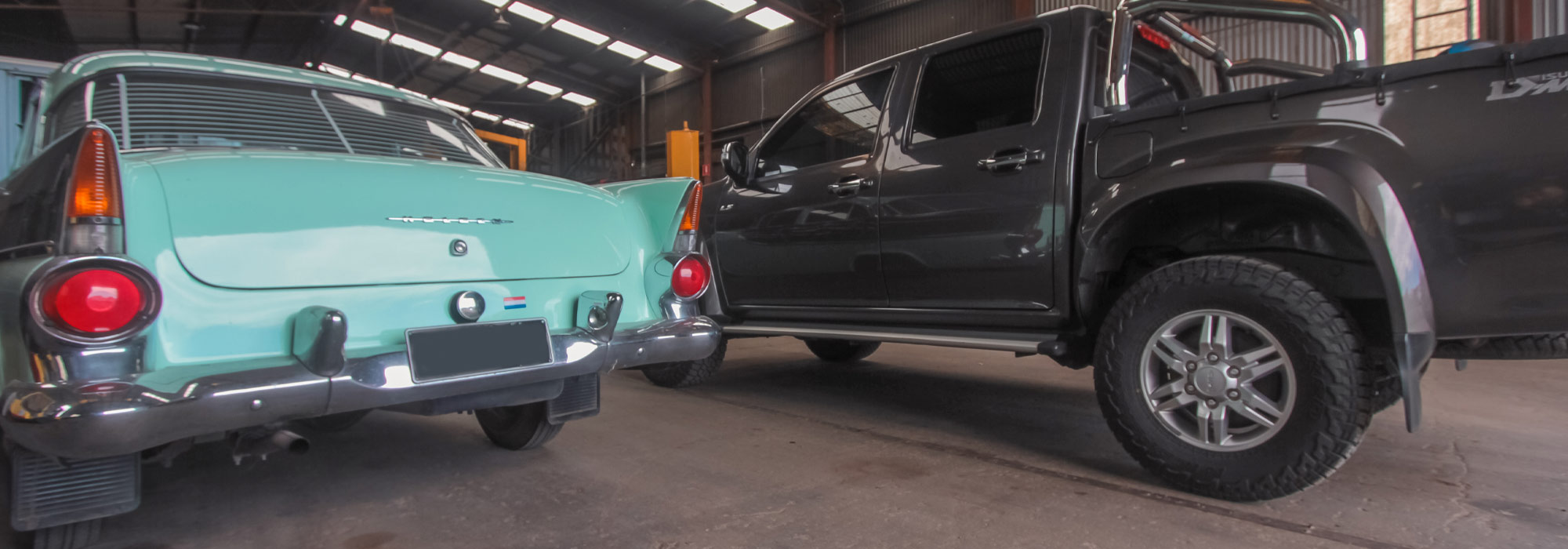 Photo of the rear of an aqua coloured classic car and beside it a twin cab ute that is charcoal colour.