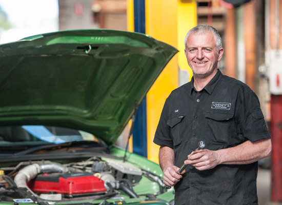 Photo of mechanic Ian McEwan wearing black shirt holding a tool, behind him is a green car with the bonnet up and engine showing.